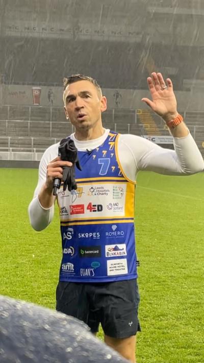 Kevin Sinfield with microphone and gloves in one hand raises his other hand during a speech at Headingley Stadium.