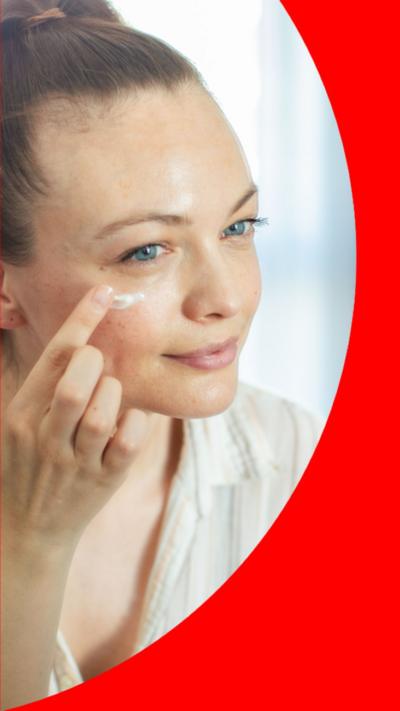 A woman with dark hair pulled back in a ponytail applies a facial skincare product in the mirror.