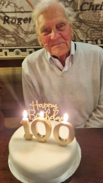 A man looking at the camera with a cake in front of him. The cake has candles which depict 100 and say Happy Birthday.