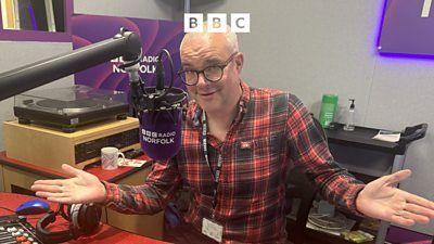 Chris Goreham sits in front of a bbc radio norfolk purple branded microphone with his arms out to the side and a quizzical expression on his face. he wears a plaid shirt, glasses on his face and a bbc lanyard