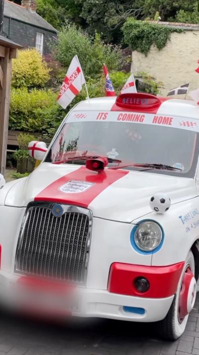 A white and red car which has "its coming home" written in red letters. There are english flags and footballs over the car. The number plate is blurred.