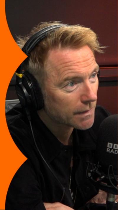 A man with short blond hair sits in front of a microphone and talks to someone off-camera
