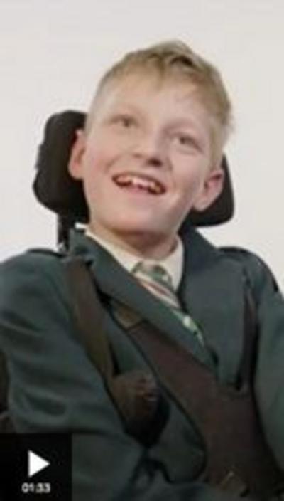A young boy sits in a wheelchair, smiling and looking just above the camera. He has short blonde hair and is wearing a dark green school uniform