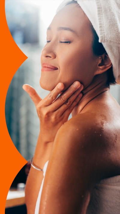 A woman stands in a bathroom with her eyes closed, touching her face. Her hair and body are wrapped in towels