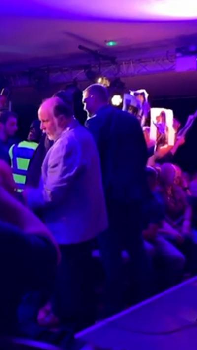 John Swinney sits on a stage  while pro-palestinian protestors in the crowd stand and hold placards.