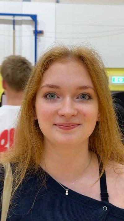 A red haired teenage girl looks into the camera with a smile. She wears an off the shoulder black top.