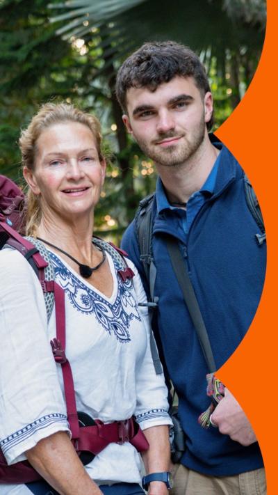 Caroline and Tom are both wearing large backpacks. They are standing together in front of shrubbery.