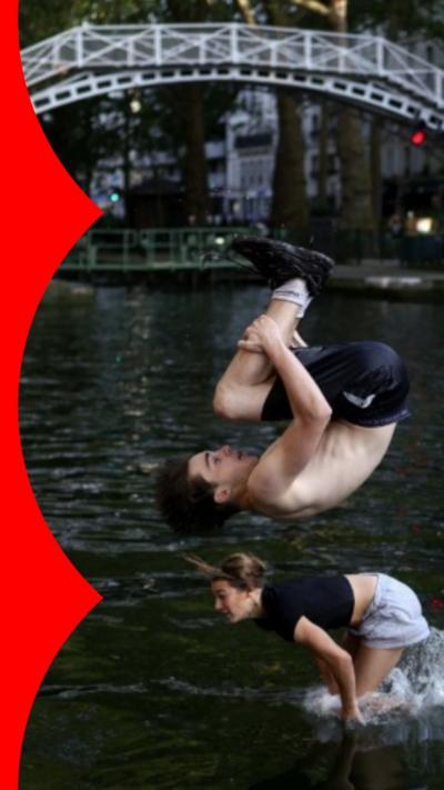 A topless young man with brown hair wearing black shorts and trainers somersaults into the water at Canal St Martin.