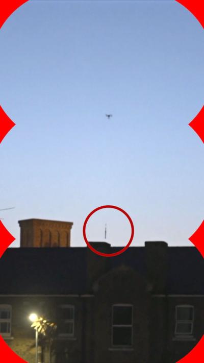 A drone is seen flying in a blue sky above HMP Wandsworth - it is dropping what appears to be a long packaged into the prison and the object being dropped is has a red circle on it to make it more obvious