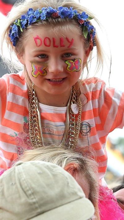 A young girl wears a flower crown and has the word "Dolly" written on her forehead.