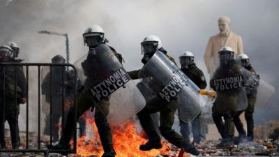 Two Greek police officers in riot armour holding shields with a fire, tear gas and more officers behind them