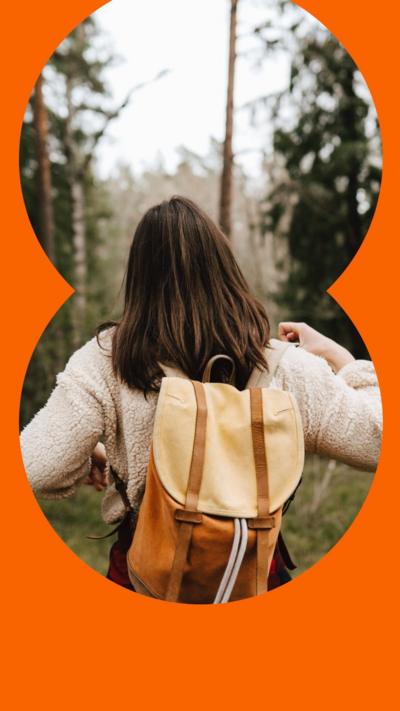 A woman walks in a forest - she has long brown hair and wears a woolly jacket and backpack 