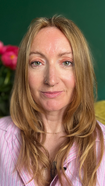 A woman with blonde hair and a nose ring looking down the barrel. There is a pink flower behind her