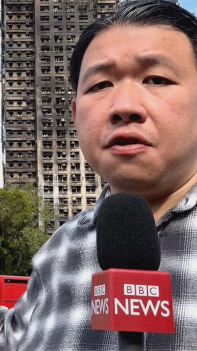 Reporter Martin Yep wearing chequered shirt holding BBC News-branded microphone standing in front of burned out building