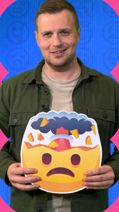 A man smiles at the camera and holds an emoji in his hands