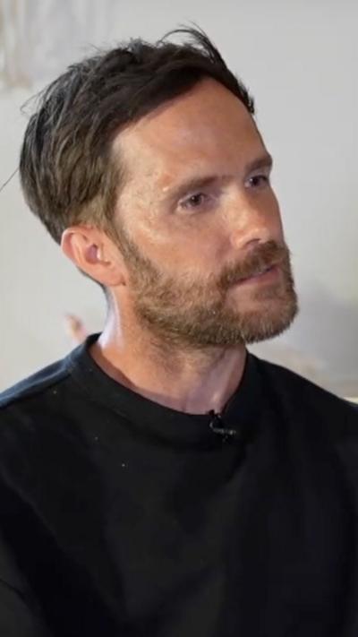 Man in black jumper looks ahead. He has dark hair which is swept from left to right and he has stubble. 