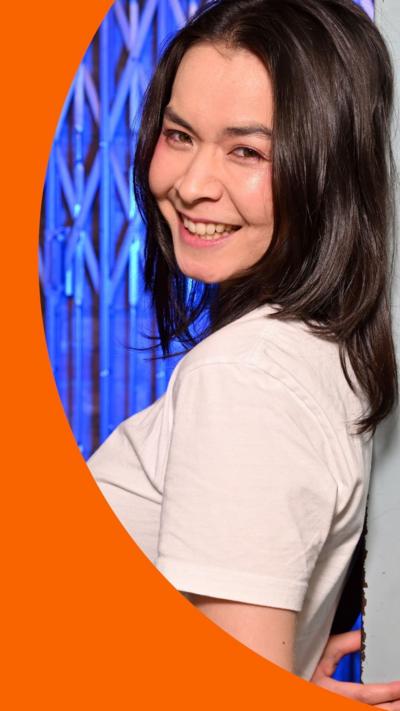 Mitski wears a white T-shirt and stands smiling with her back against a metal folding door.