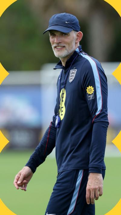 Thomas Tuchel wears a blue cap and England training tracksuit. He looks at the camera and smiles.