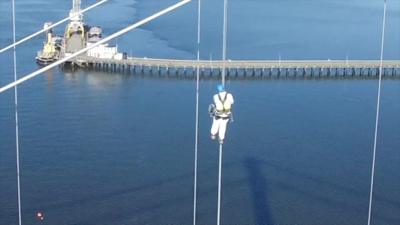 A man in white clothes and a blue helmet. He is abseiling on the Tamar Bridge. The river is blue. 