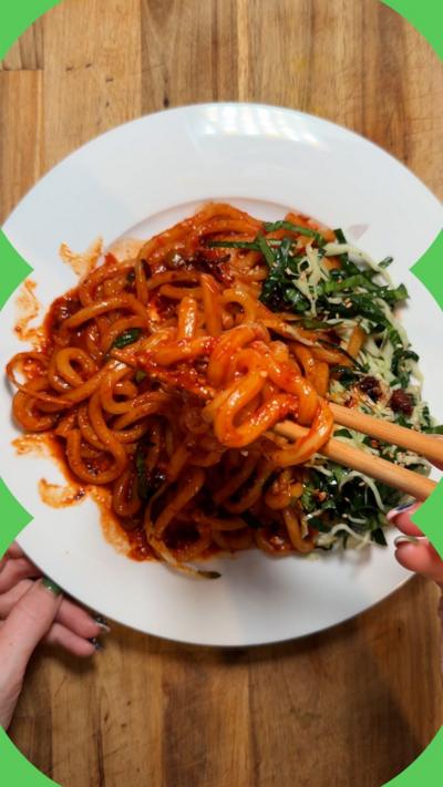 A white plate with noodles in a tomato sauce being picked up by a pair of chopsticks. Two hands are seen either side.