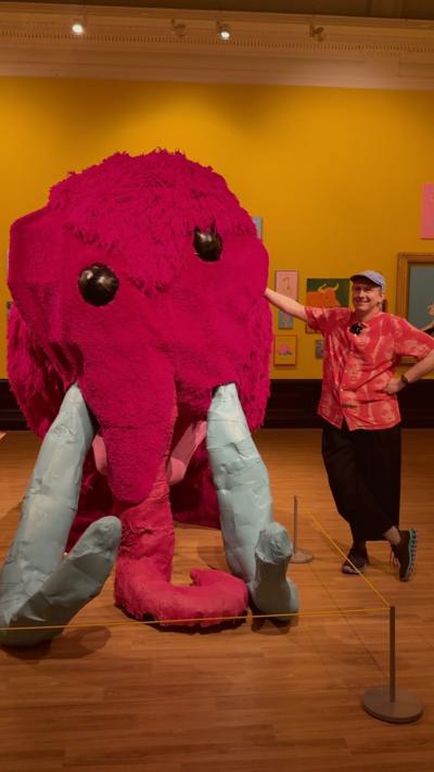 a man in an orange and red shirt with cap and black trousers next to a pink mammoth sculpture