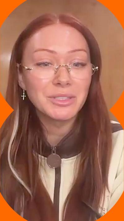 Elz The Witch has long red hair and is wearing rimless glasses and a cream jacket.