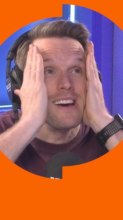 Greg Foot clasps his head in his hands and looks worried. He is wearing a brown T-shirt and is sat behind a Radio 4 mic.