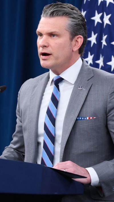 Pete Hegseth wears a grey suit and striped tie while speaking behind a podium.