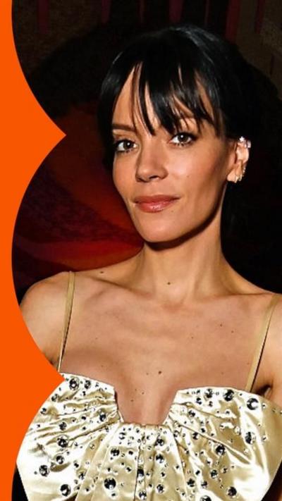 Lily Allen, looking to camera, wearing a gold strap dress, with her hand on a wooden pole.