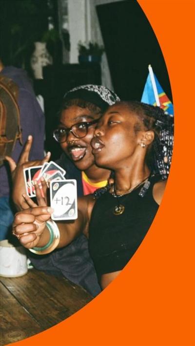 Two young women playing uno, one of them is showing a card with '+12' written on it