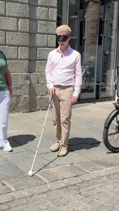 Man in a pink shirt and cream trousers with a black blindfold and holding a white cane