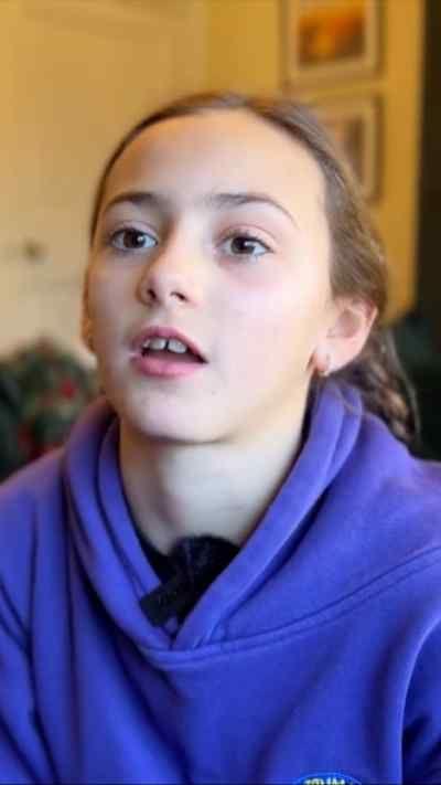 A young girl with brown hair tied back looks at the camera as she wears a purple hoody.