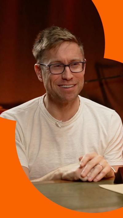 Russell Howard sits behind a desk with his hands on the table. He has short blond hair and wears a white T-shirt