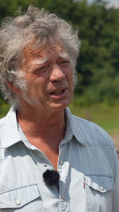 Guy Singh-Watson is wearing a blue short-sleeved shirt as he stands in the bright sunshine in a dry reservoir on a farm at Riverford Organic Farmers in Devon.