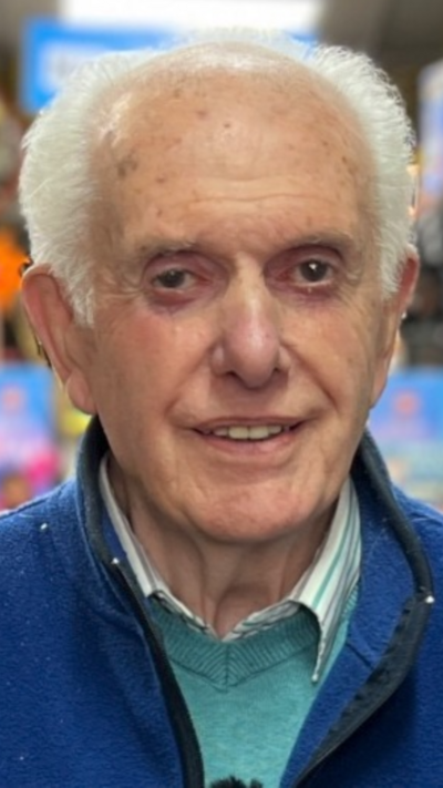 An elderly man with grey hair and a blue jacket on. He is smiling at the camera
