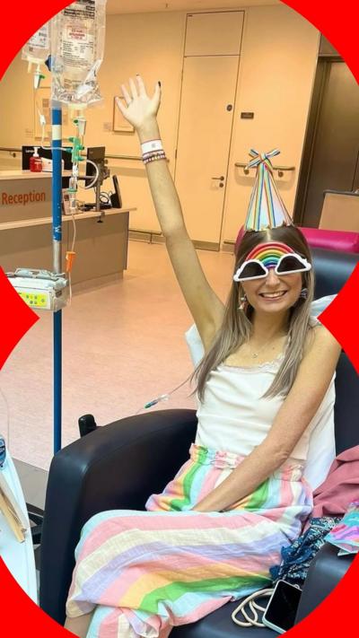 Florence Moffatt is sitting down wearing rainbow sunglasses and a stripy party hat. She is connected to a drop and has her right hand in the air. She is smiling. 