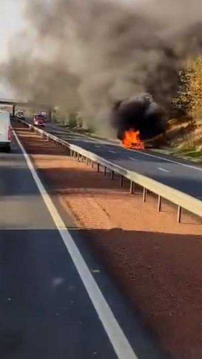 Dashcam footage of a car in flames at the side of a motorway with black and grey smoke.