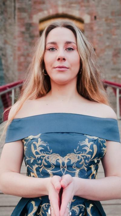 A woman with long blonde hair in a blue shoulderless dress stood in front of a castle entrance 