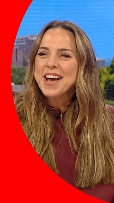 Melanie C wearing a burgundy hoody, smiling, with long brown wavy hair.