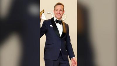 A man wearing a black tuxedo is holding up an Oscars trophy