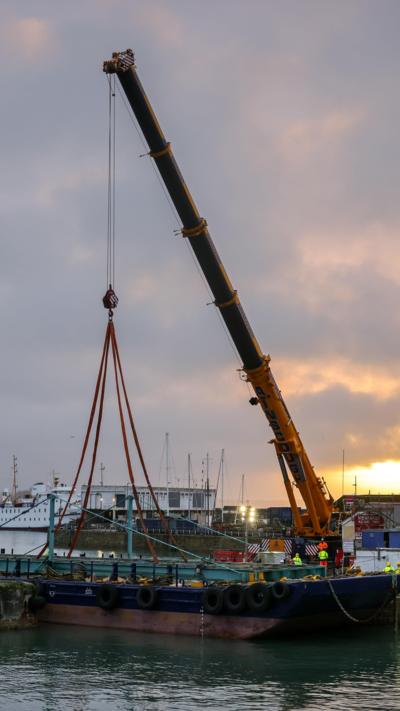 A bridge being held up by a crane just about a boat. The boat is in a harbour and there are people in high visibility clothing. The sea is blue. The sky is blue and cloudy. 
