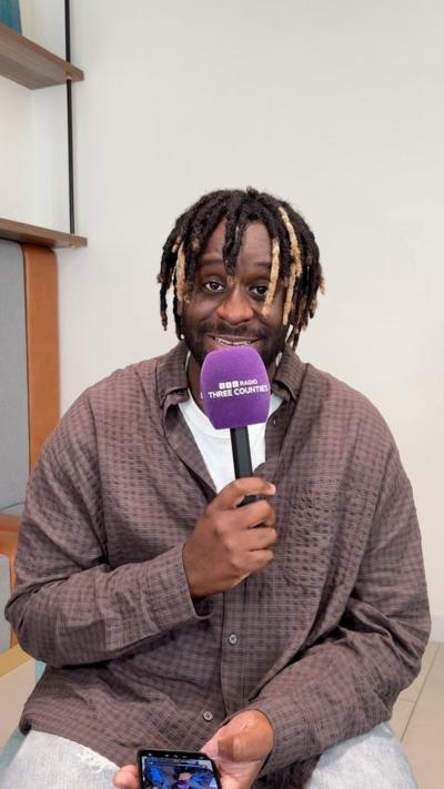 Myles Smith is smiling, staring at the camera. He is hold a BBC Three Counties Radio branded purple mic. He is wearing a checkered brown shirt.