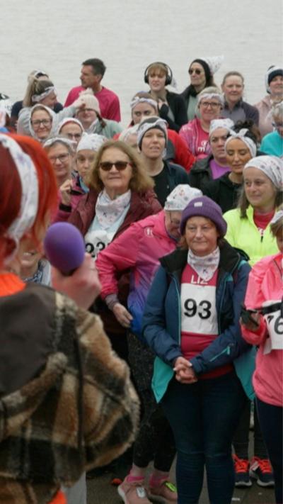 A large group of participants wearing bright running and support vests gather near a waterfront. The crowd appears engaged, many holding phones or clapping, creating an energetic and communal atmosphere.