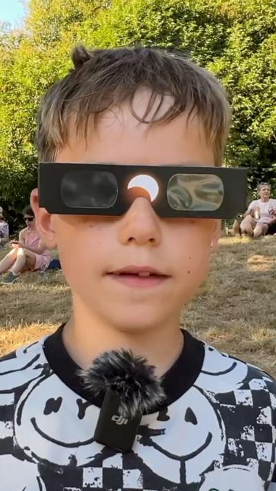 Boy with brown hair wearing eclipse glasses while stood in a field with trees in the background