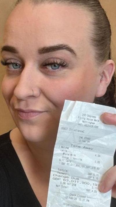 Young woman with blonde/brown curly hair smiling at the camera with a grey lanyard on. She's wearing a green and pink polka dot top with a high ruched collar. Behind her is a tissue dispenser and an orange medical bin. The wall behind her is white, and there is half of a blue doorframe to the left of her. 