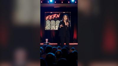 Fern Brady is stood on a stage, speaking to an audience, with her name in lights behind her