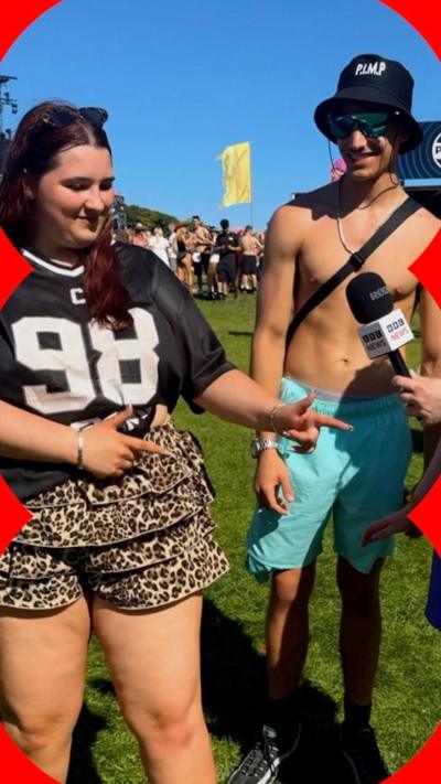 A woman and man smiling at TRNSMT festival - the woman is posing with index fingers pointing left, the man has his top off.