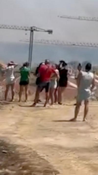 People stand on a sandy hillside as they look at wildfire smoke