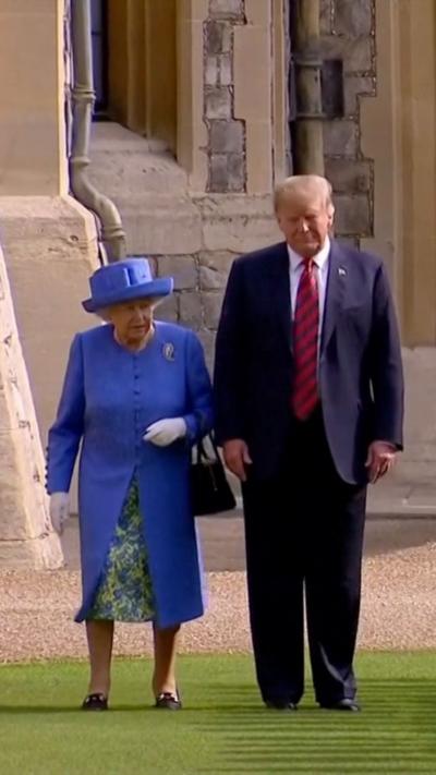 The Queen wears a blue suit and hat and walks next to Donald Trump who wears a suit and red striped tie