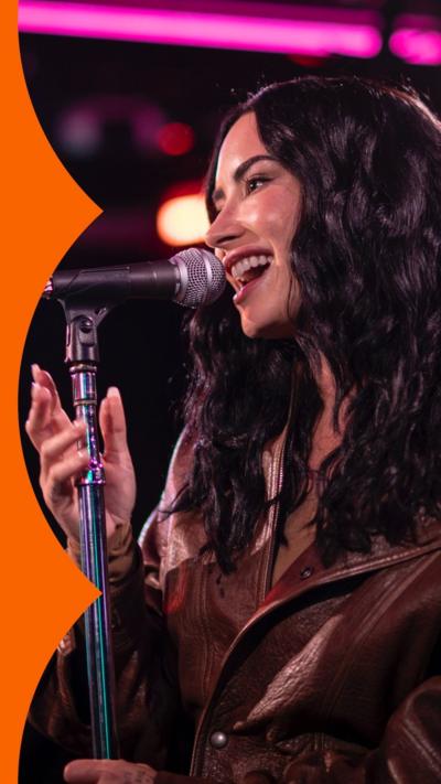 Demi Lovato sings into a mic in the Radio 1 Live Lounge. She has wavy dark hair and wears a brown jacket